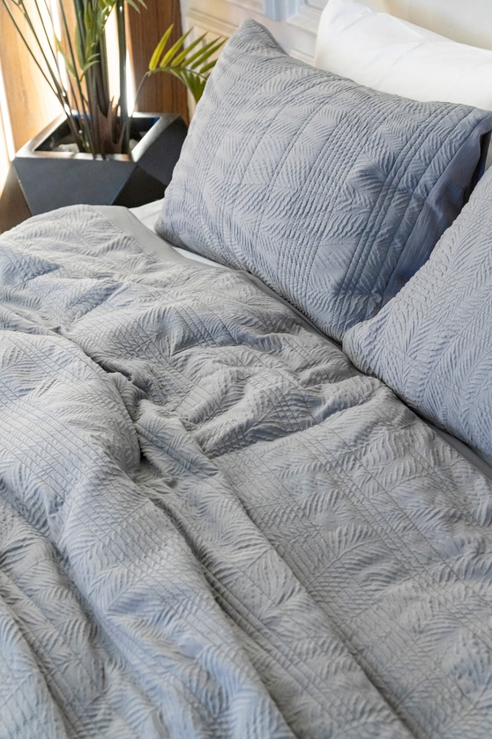 A close-up of a textured gray quilted comforter and matching pillowcases on a bed, with a potted plant visible in the softly lit background.