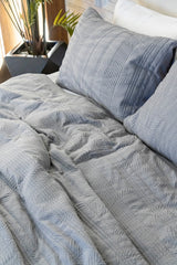 A close-up of a textured gray quilted comforter and matching pillowcases on a bed, with a potted plant visible in the softly lit background.