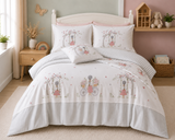 A children's bedding set styled on a bed in a nursery, featuring a white and light gray striped design with pastel pink and green illustrations of girls on bicycles and trees.