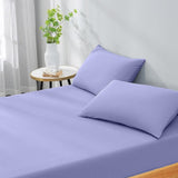 Purple bed sheets and pillows on a bed with a side table and plant in the background.